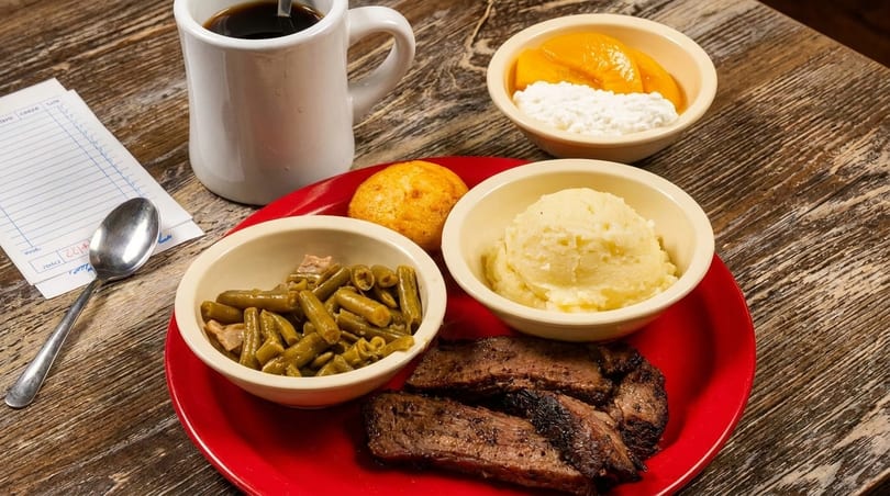 A red plate with sliced brisket, green beans, mashed potatoes, a corn muffin, and coffee.