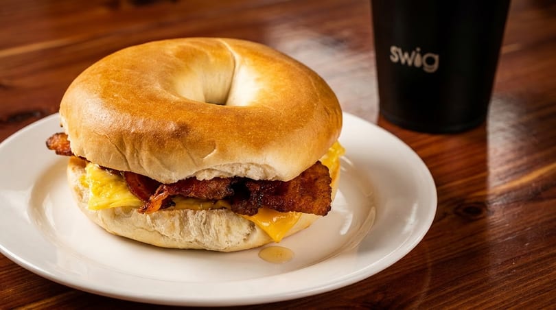A bacon, egg, and cheese bagel on a white plate next to a black Swig cup.