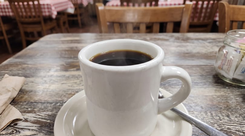Steaming hot coffee in a white mug on a rustic wooden table with a sugar caddy.