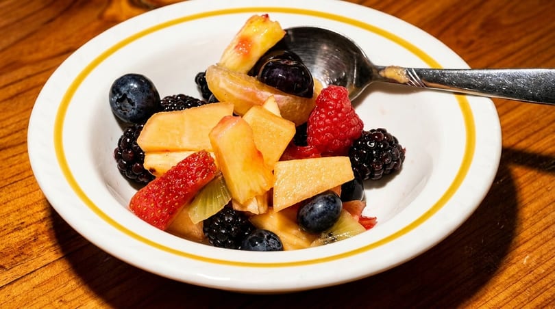 A white bowl with a yellow rim filled with mixed fruit salad and a spoon on a wooden table.